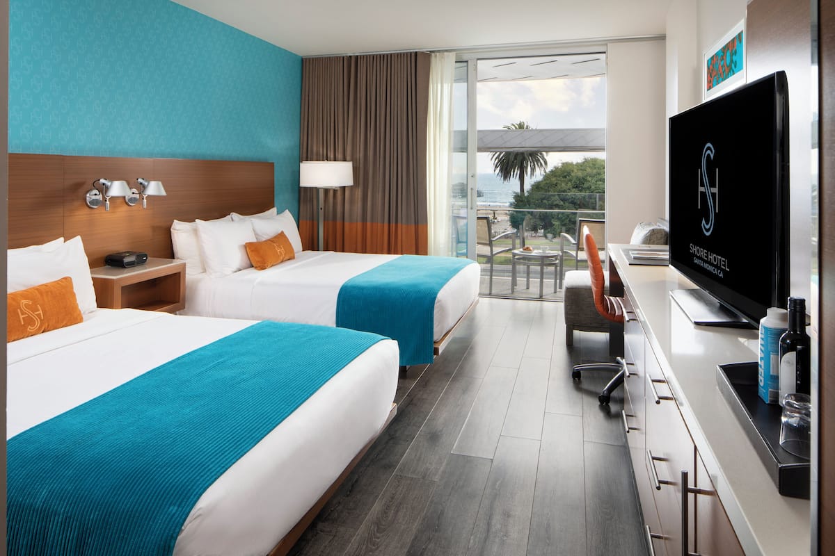 Habitación de hotel con dos camas, televisión, escritorio y balcón con vistas.