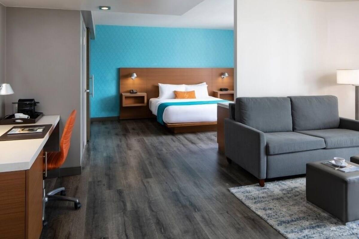 Habitación de hotel con cama, sofá gris, escritorio con silla y una mesita de noche con lámpara.