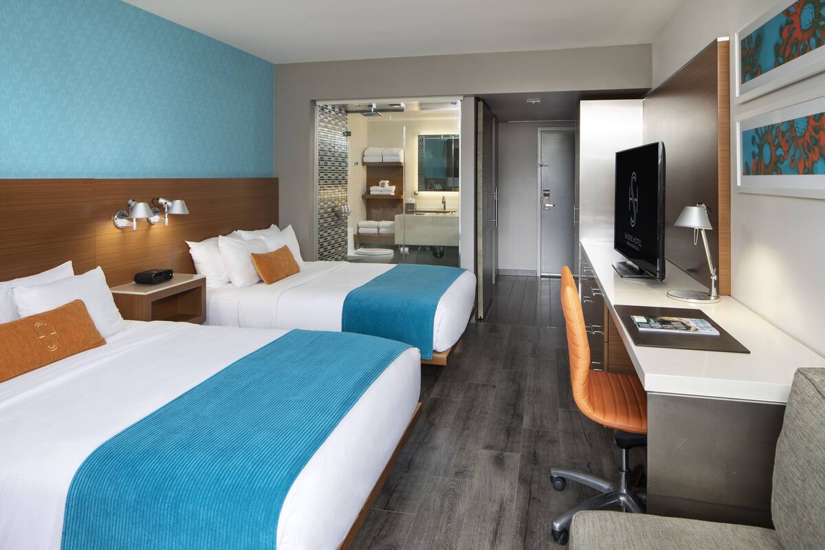 Habitación de hotel con dos camas, un escritorio, una silla, un televisor y un baño visible a través de una puerta abierta.