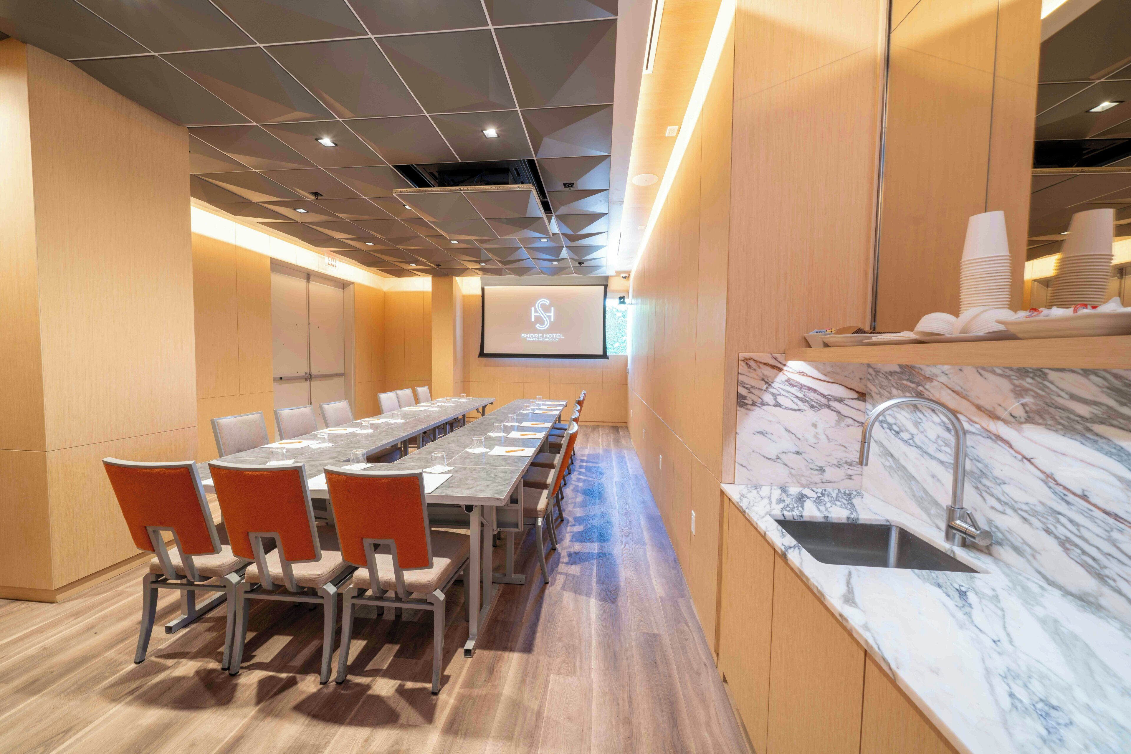 A conference room with a long table, chairs, a projector screen, and a marble countertop with a sink.