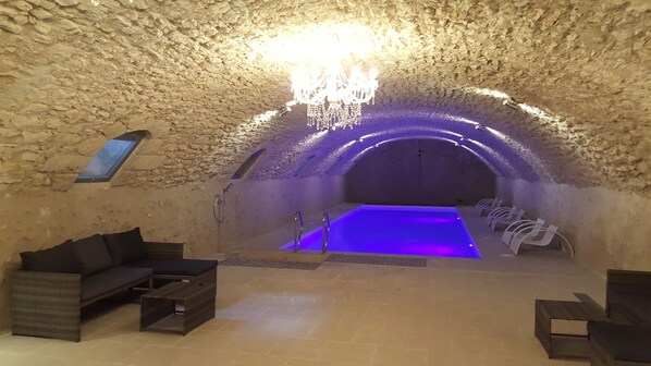 Una habitación subterránea con techo de piedra, una piscina iluminada en azul, un sofá y una araña.