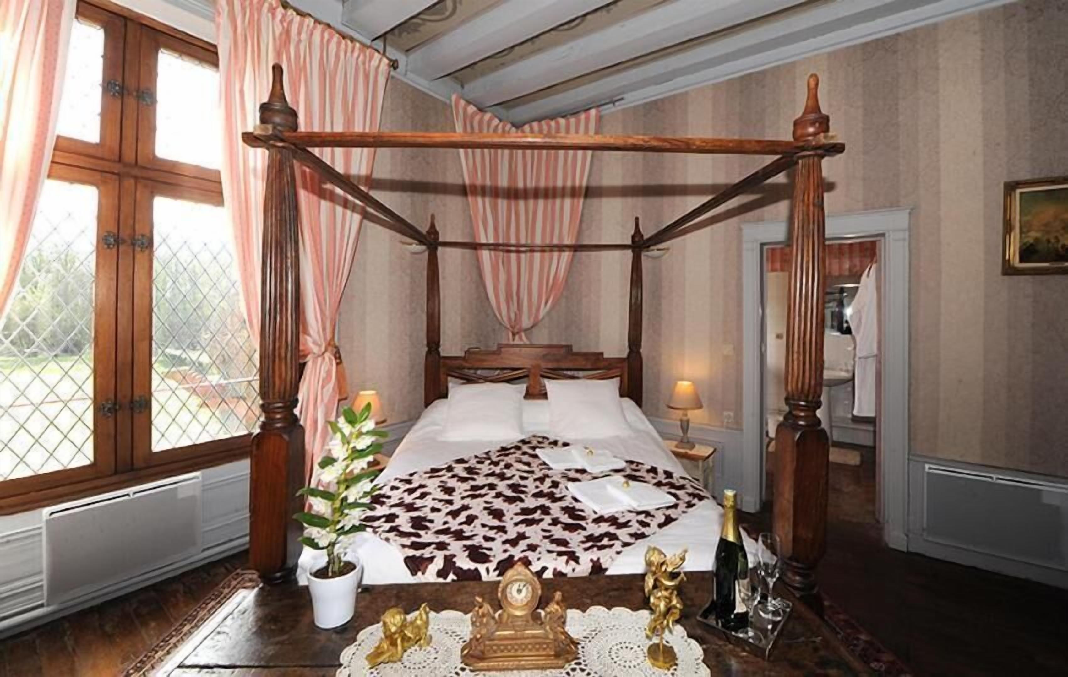 A four-poster bed with a floral bedspread, a potted plant, and a small table with a bottle and glasses.