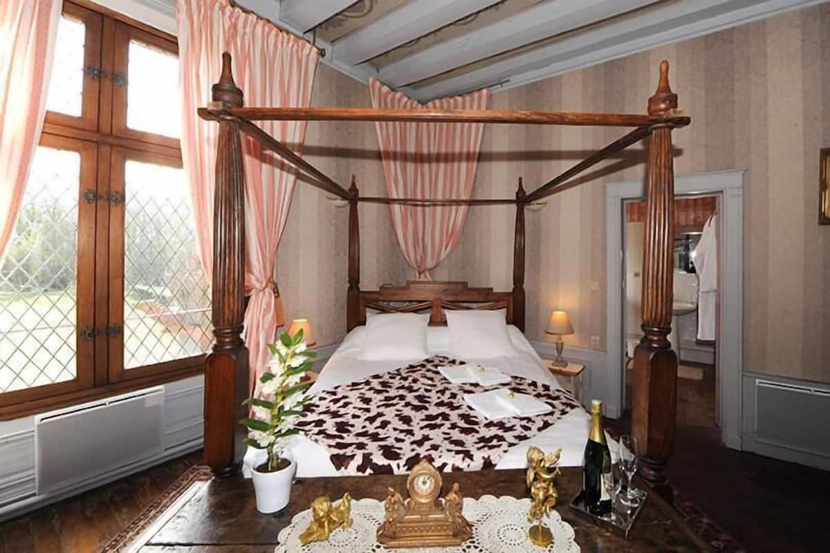 A four-poster bed with a floral bedspread, a potted plant, and a small table with a bottle and glasses.