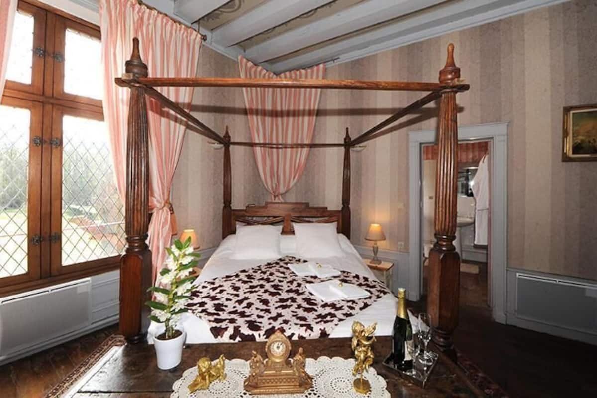 A four-poster bed with a floral bedspread, a potted plant, and a small table with a bottle and glasses.