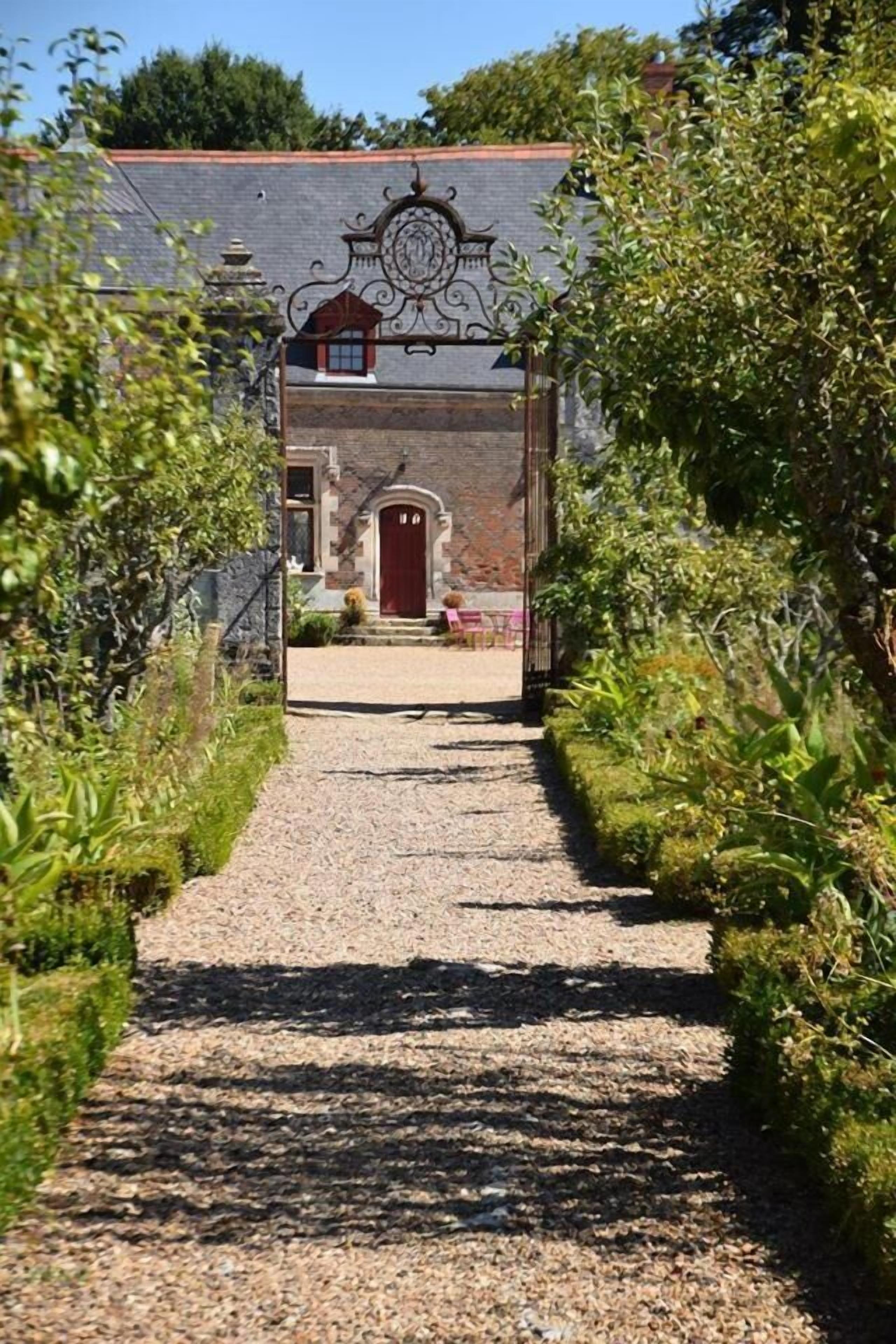 A gravel pathway leading to a stone building with a red door, surrounded by greenery.