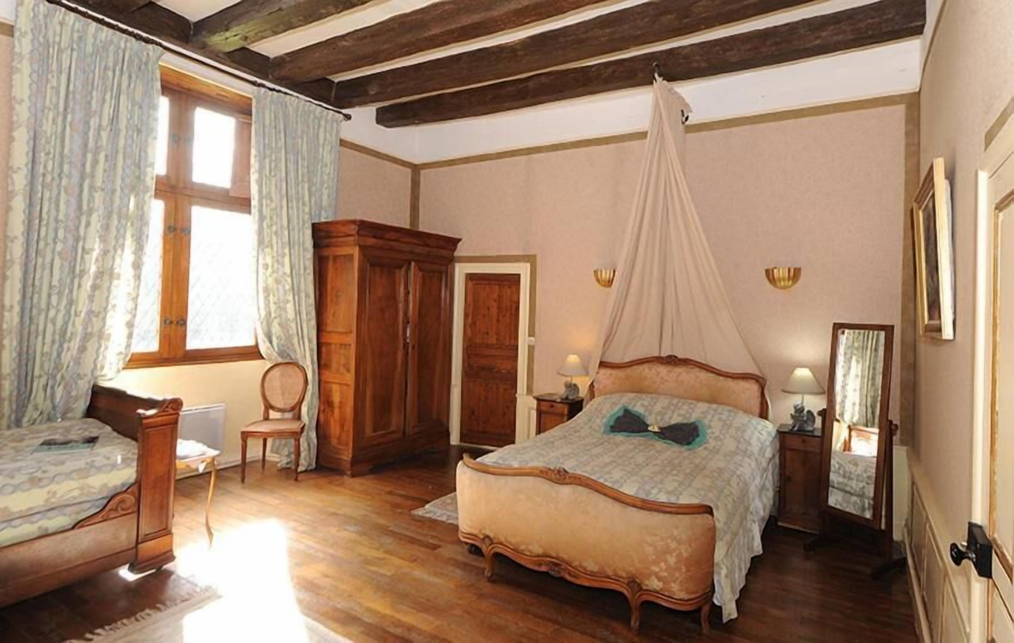 A bedroom with a large bed, a smaller bed, a wooden wardrobe, a chair, a bedside table with a lamp, and a canopy bed.