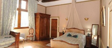 A bedroom with a large bed, a smaller bed, a wooden wardrobe, a chair, a bedside table with a lamp, and a canopy bed.