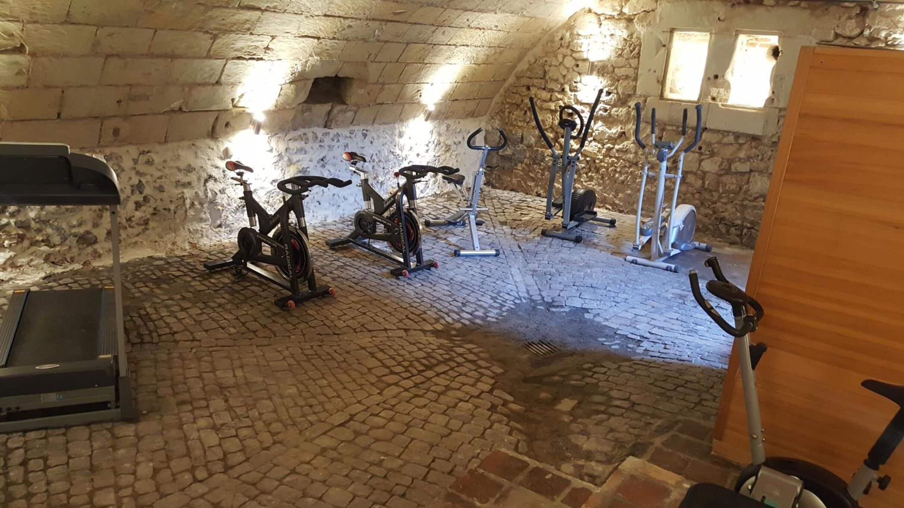 Una habitación con suelo de ladrillo y paredes de piedra, que contiene varias bicicletas estáticas y una cinta de correr.