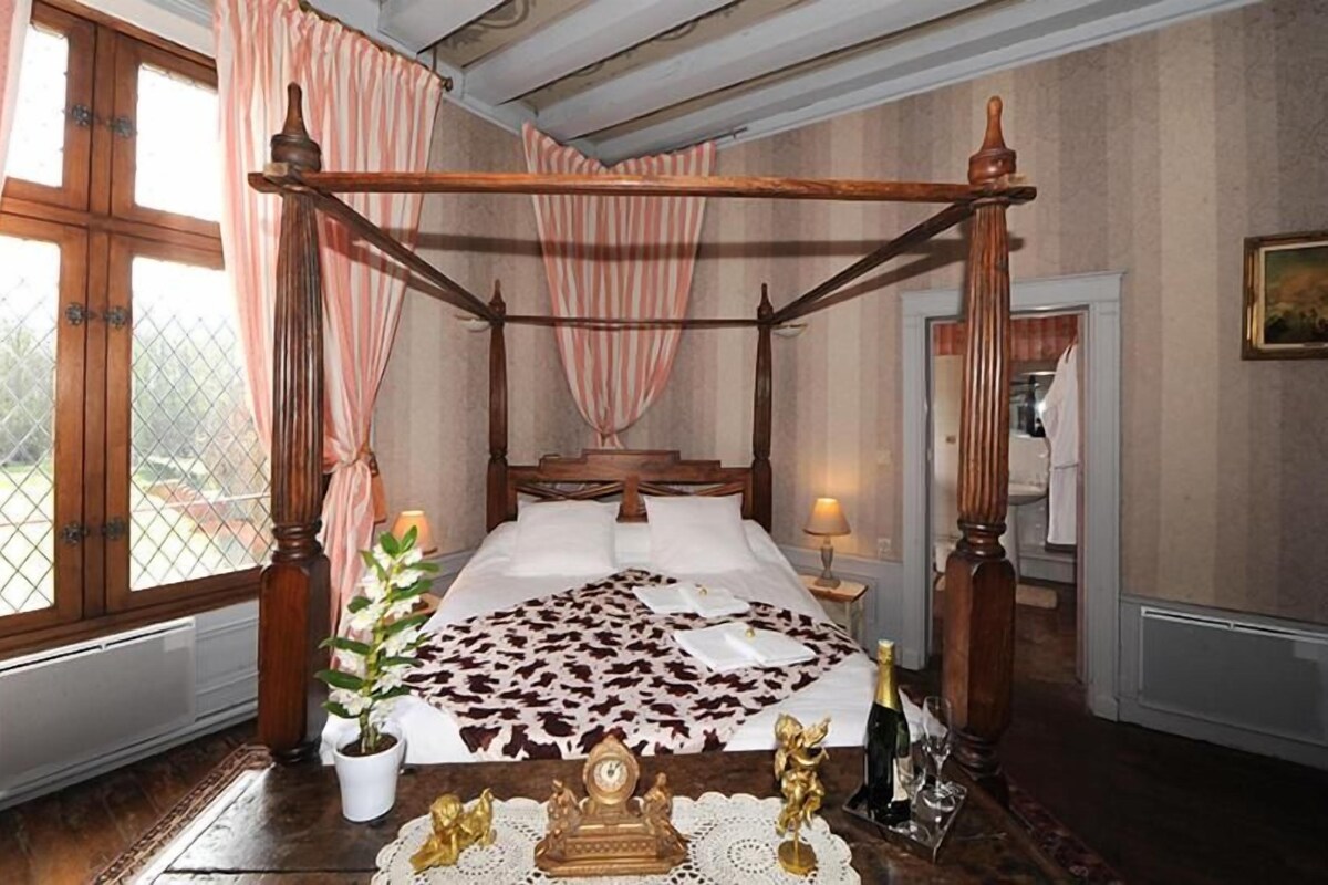 A four-poster bed with a floral bedspread, a potted plant, and a small table with a bottle and glasses.