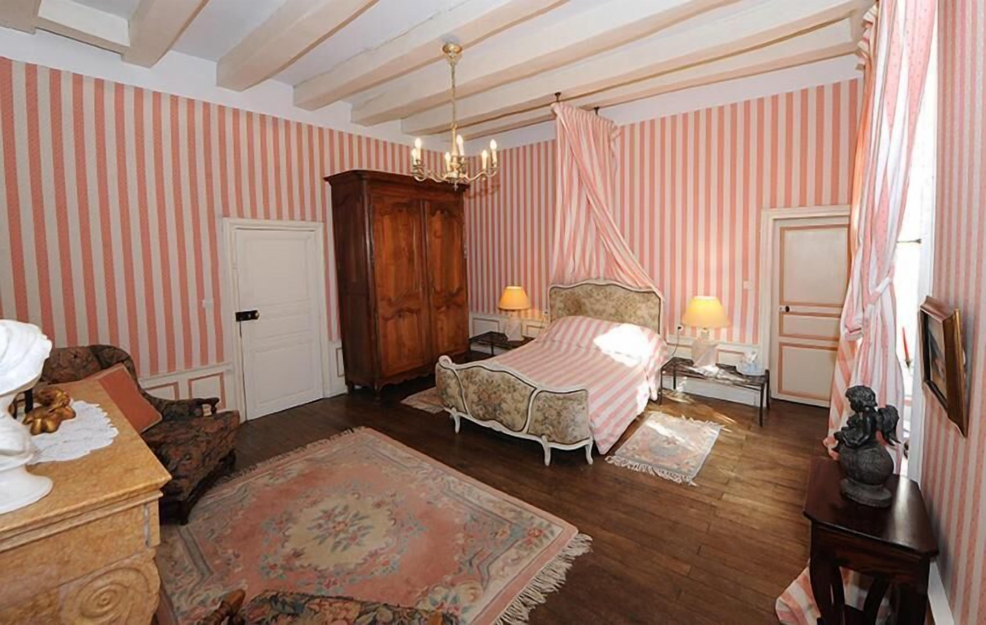 A room with striped wallpaper, a bed with a canopy, a chandelier, a wooden wardrobe, and a desk with a lamp.