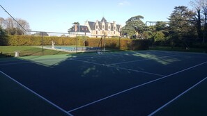 Una cancha de tenis al aire libre con red, rodeada por una cerca, y un edificio grande con una torre al fondo.