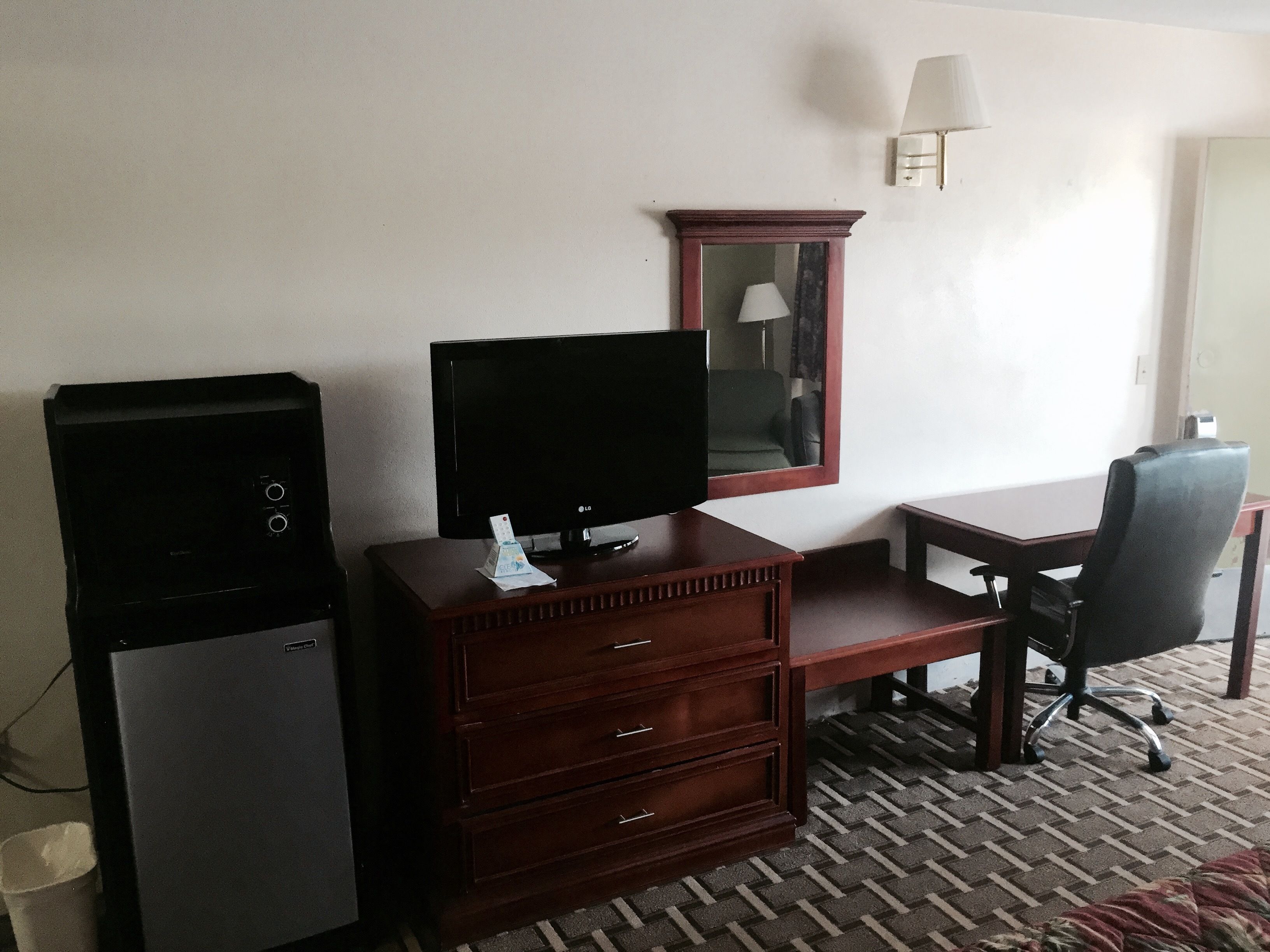A hotel room with a TV, a wooden dresser, a desk, a chair, and a lamp.
