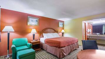 A hotel room with a bed, a green armchair, a wooden nightstand, two bedside lamps, a wooden desk with a chair, and a bathroom mirror.