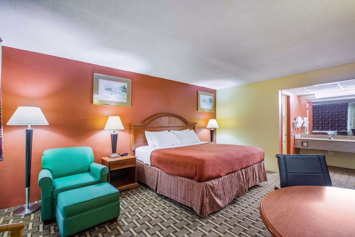 Una camera d'albergo con un letto, due lampade ai comodini, una poltrona verde, un tavolino e uno specchio sopra il lavandino.
