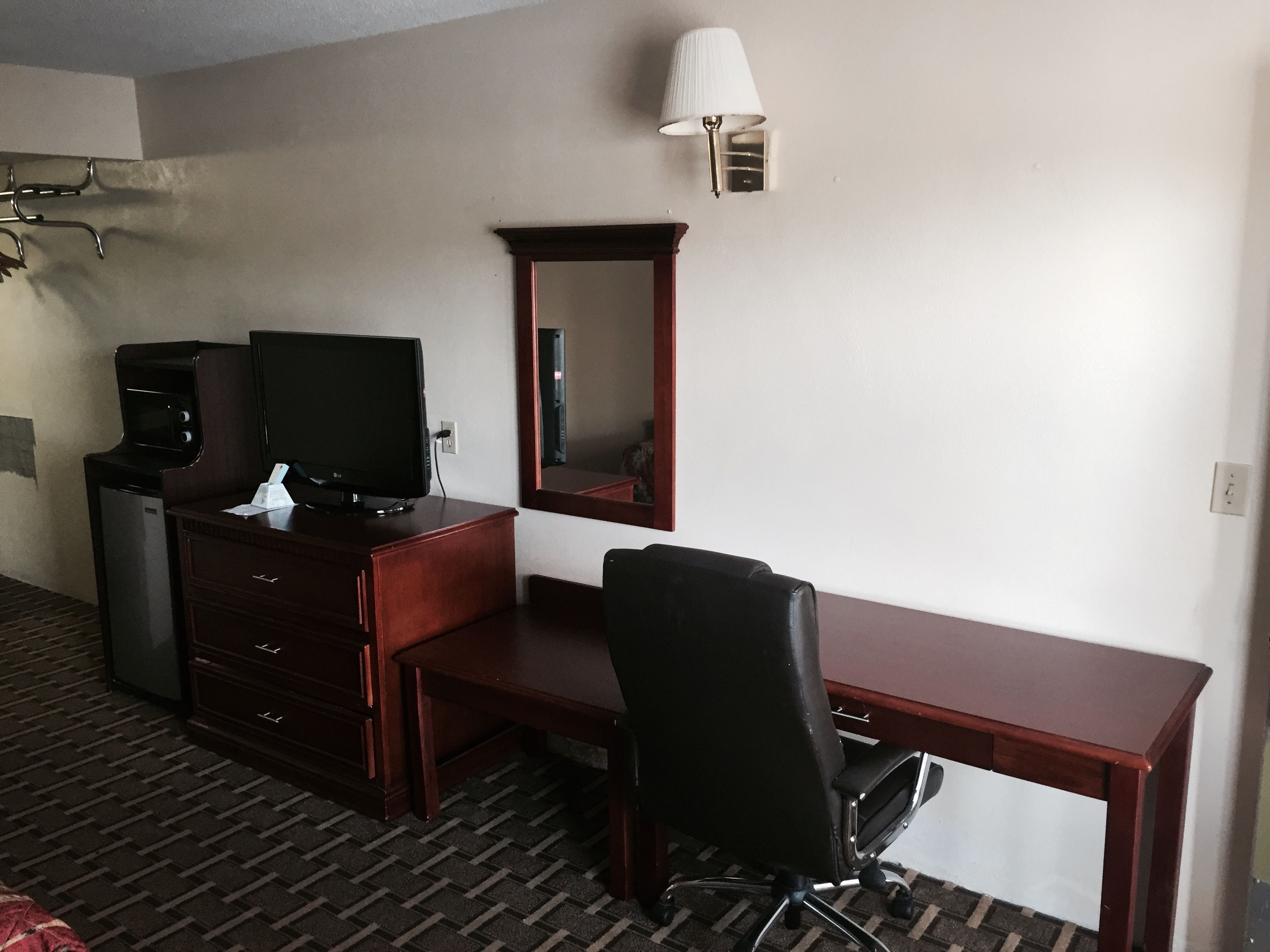A hotel room with a wooden desk, a black office chair, a television, a microwave, and a lamp.