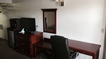 A hotel room with a wooden desk, a black office chair, a television, a microwave, and a lamp.