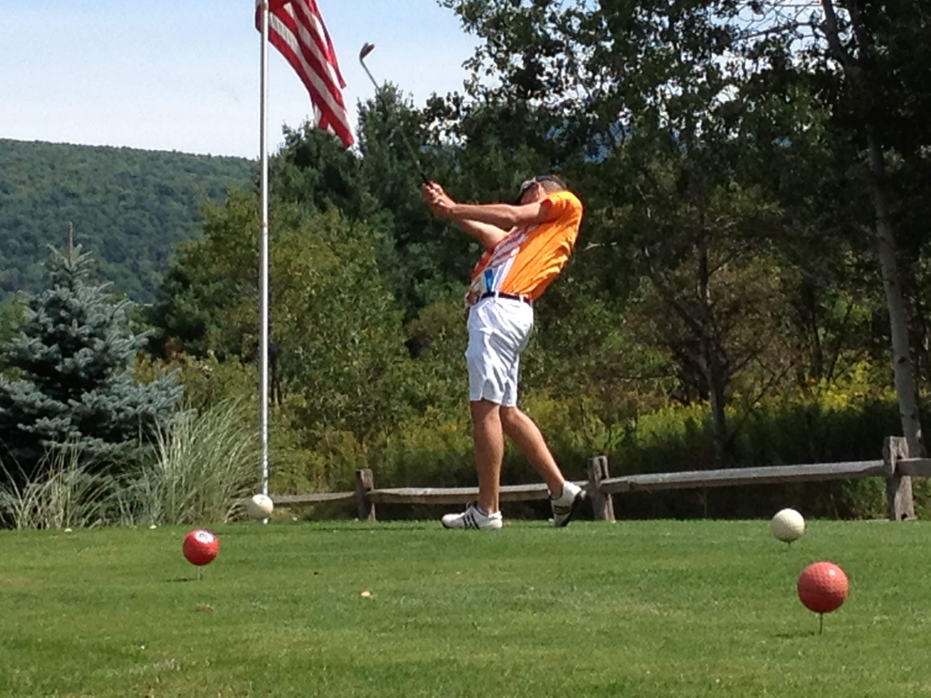Une personne jouant au golf sur un parcours avec un drapeau, des arbres et une clôture en bois en arrière-plan.