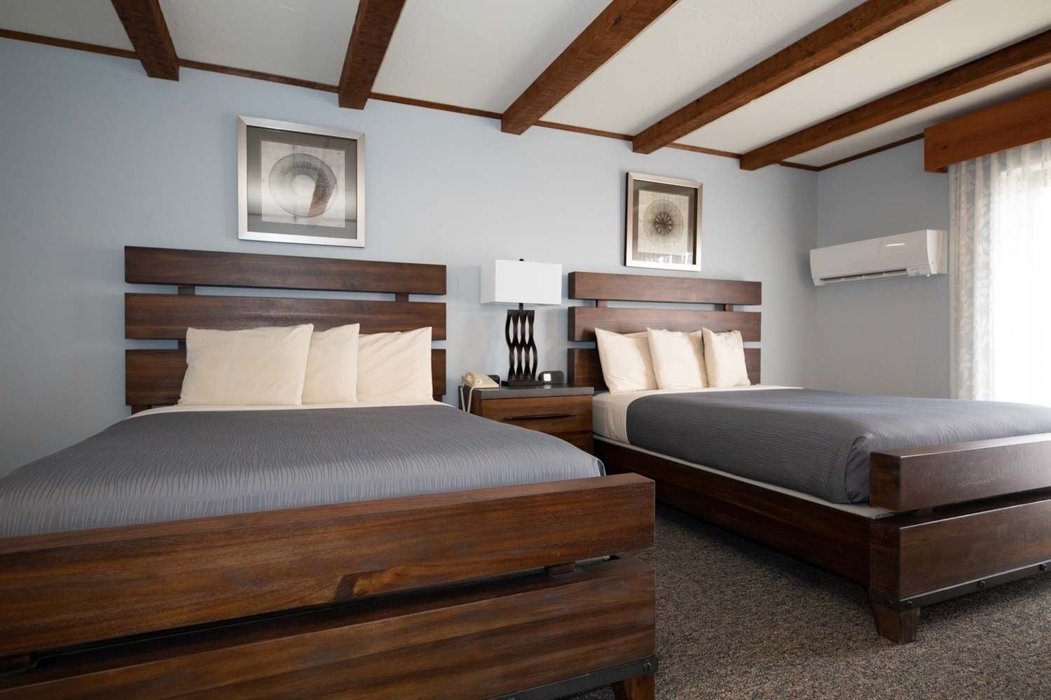 Habitación de hotel con dos camas de madera, una mesita de noche con l ámpara y obras de arte en las paredes.