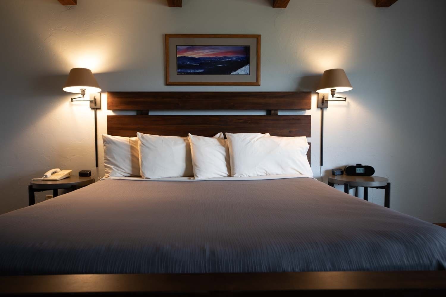 Una cama bien hecha con cabecera de madera, dos lámparas de noche, un cuadro enmarcado sobre la cabecera y una mesita de noche con un teléfono y una radio.