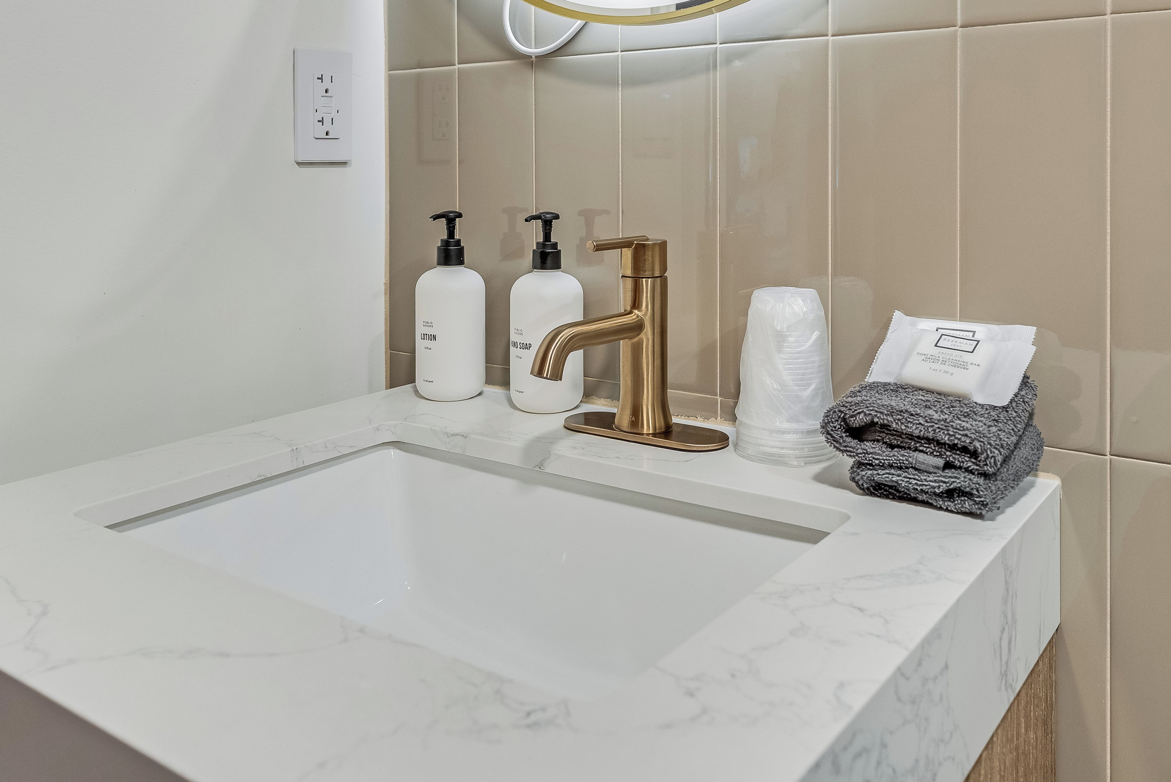 Uma pia de banheiro com bancada de mármore, uma torneira dourada e produtos de higiene pessoal, incluindo um frasco de loção e um frasco de sabonete líquido.