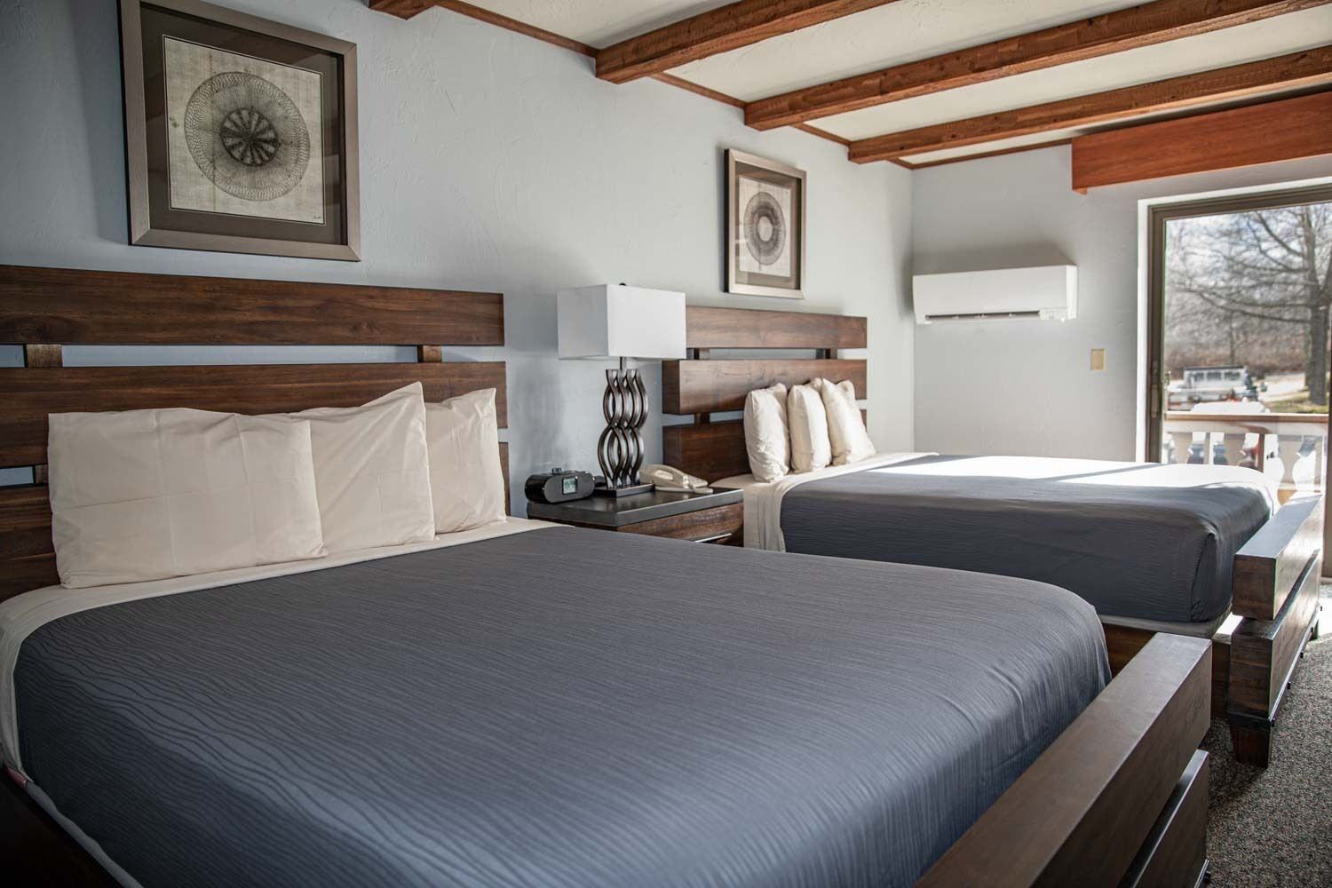 A hotel room with two beds, wooden headboards, a bedside table with a lamp, and framed artwork on the walls.