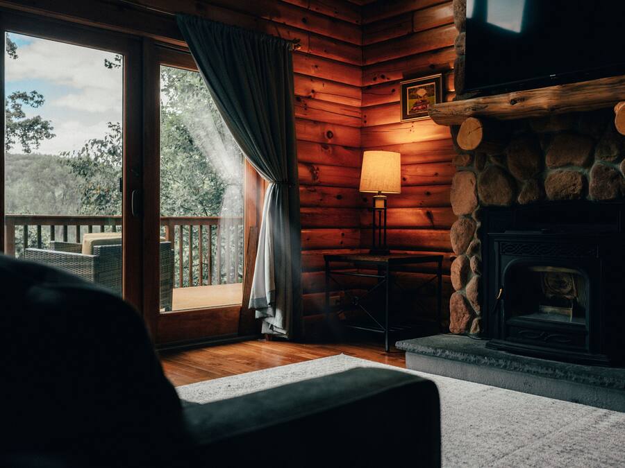 A cozy cabin interior with a stone fireplace, a wooden ceiling, and a large window with curtains.