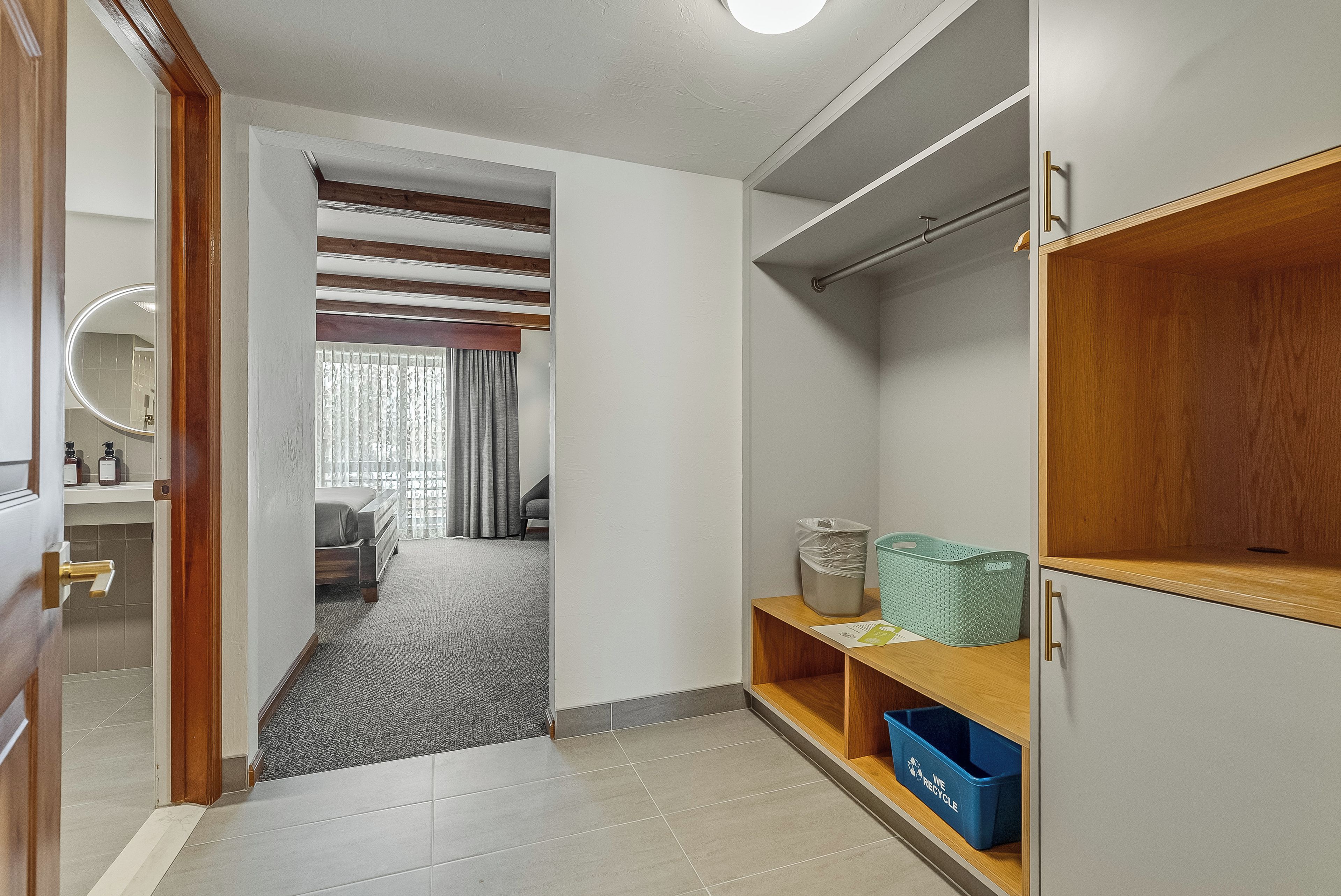 Un couloir d’hôtel avec une porte en bois, un miroir, un porte-manteau, une petite poubelle et un bac de recyclage.