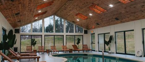 A pool area with wooden loungers, a pool, and large windows offering views of the outdoors.