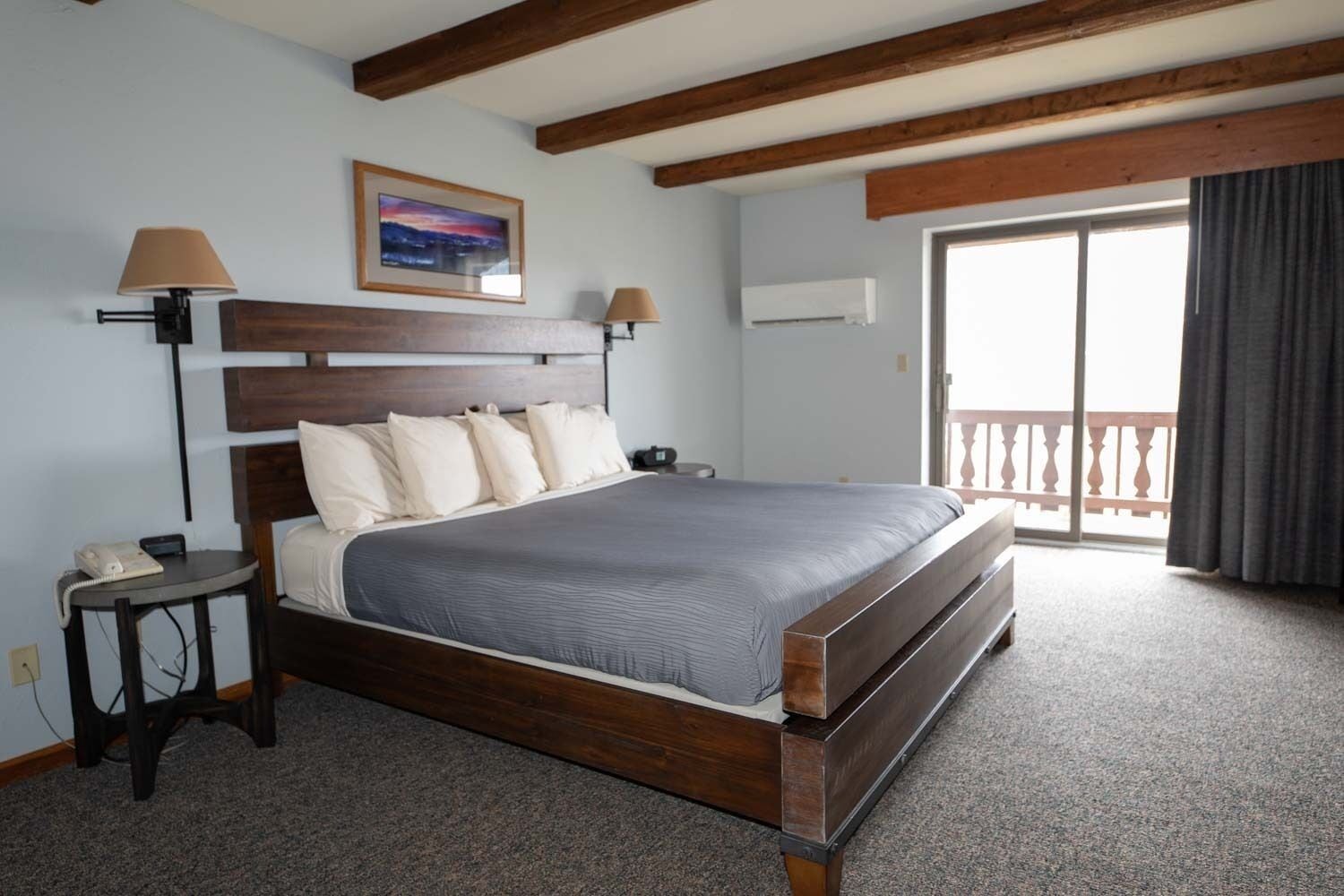 Habitación de hotel con cama de madera, una mesita de noche, una lámpara, un cuadro en la pared y un balcón con vistas.