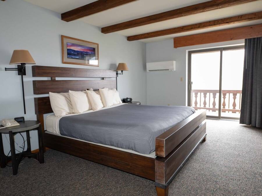 A hotel room with a wooden bed, a small bedside table, a lamp, a painting on the wall, and a balcony with a view.
