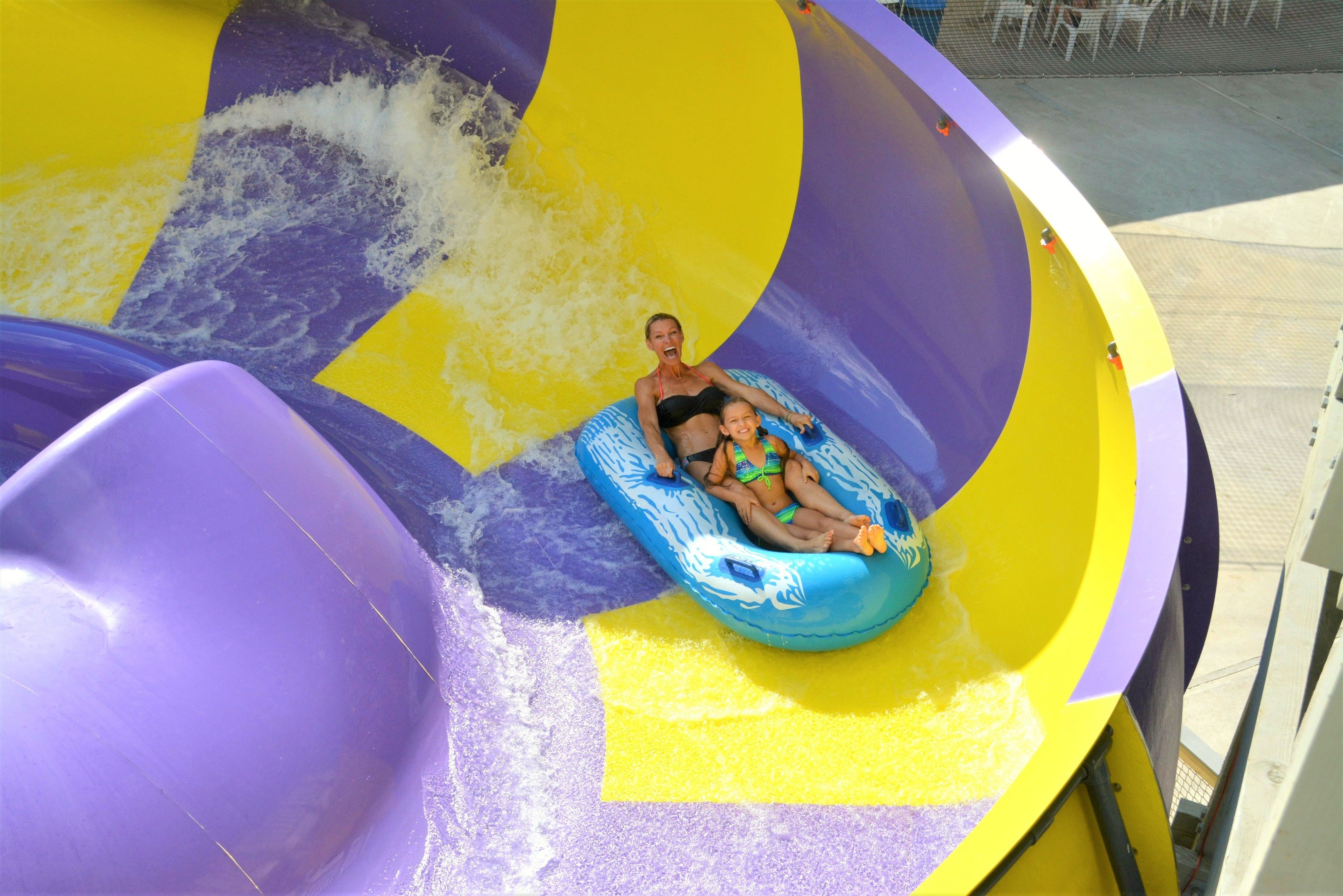 A water slide with a purple and yellow design, featuring a person and a child in a blue inflatable tube.