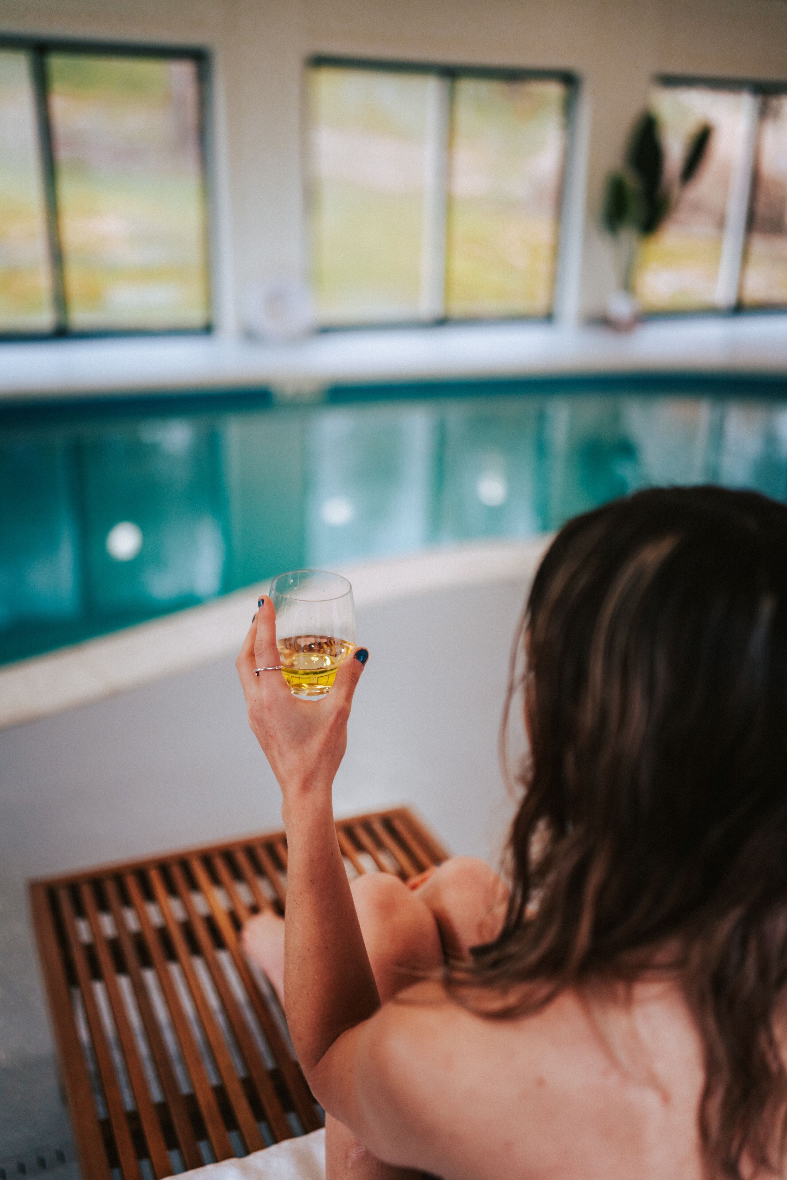 A person relaxing by a poolside with a drink, wooden slats, and a view of the outdoors through large windows.