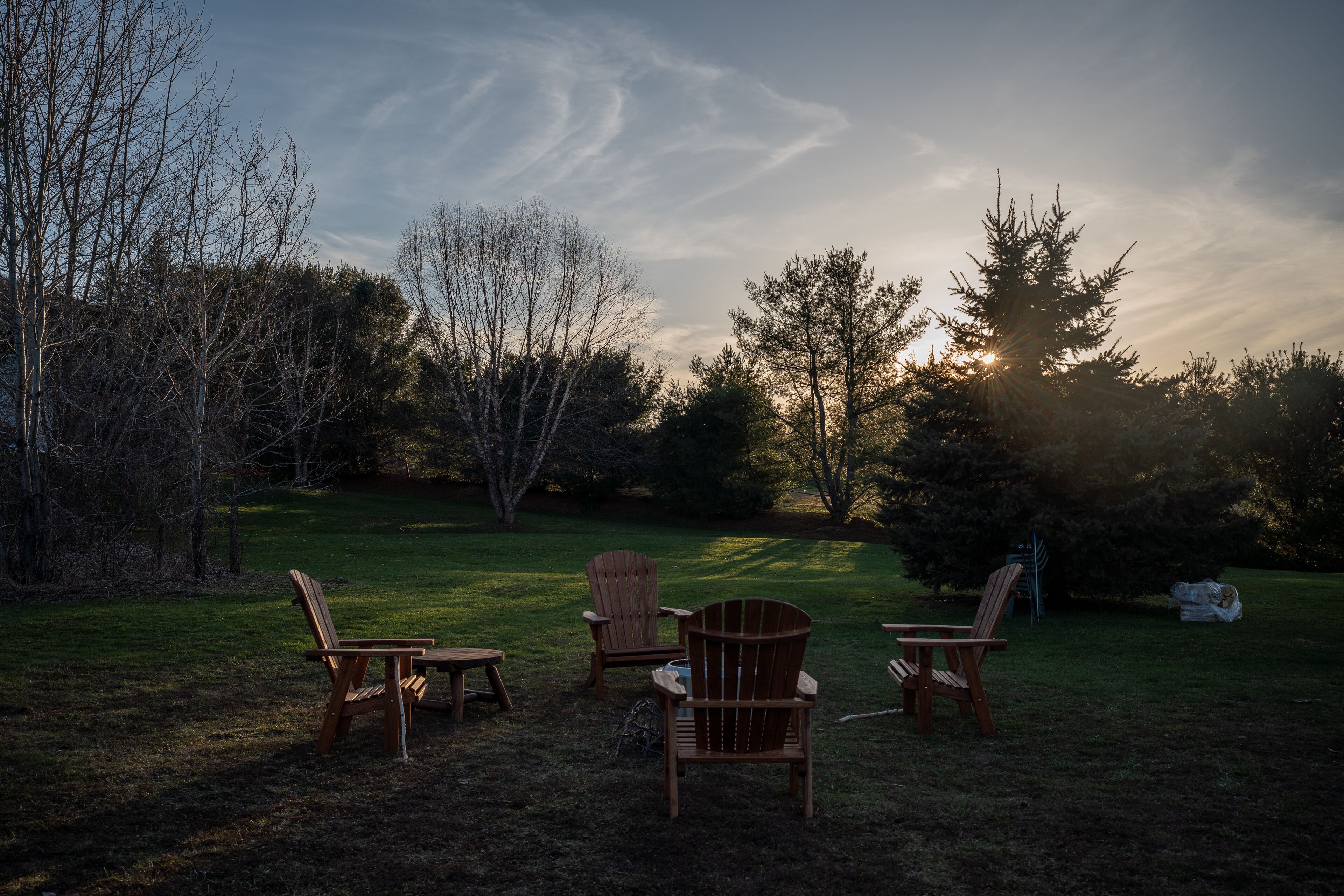 Un espacio al aire libre con césped, cuatro sillas Adirondack de madera dispuestas en semicírculo, rodeado de árboles y un cielo despejado.