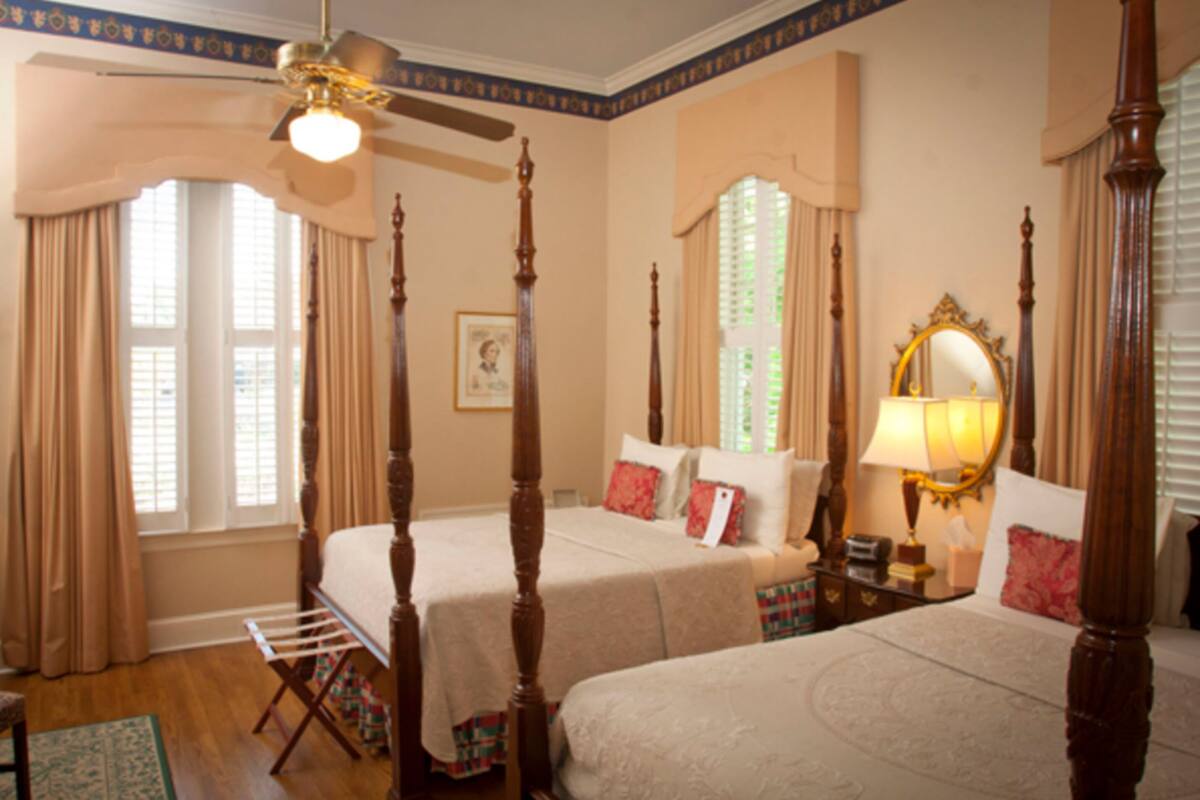 A bedroom with two beds, a ceiling fan, a window with curtains, a bedside table with a lamp, and a framed picture on the wall.
