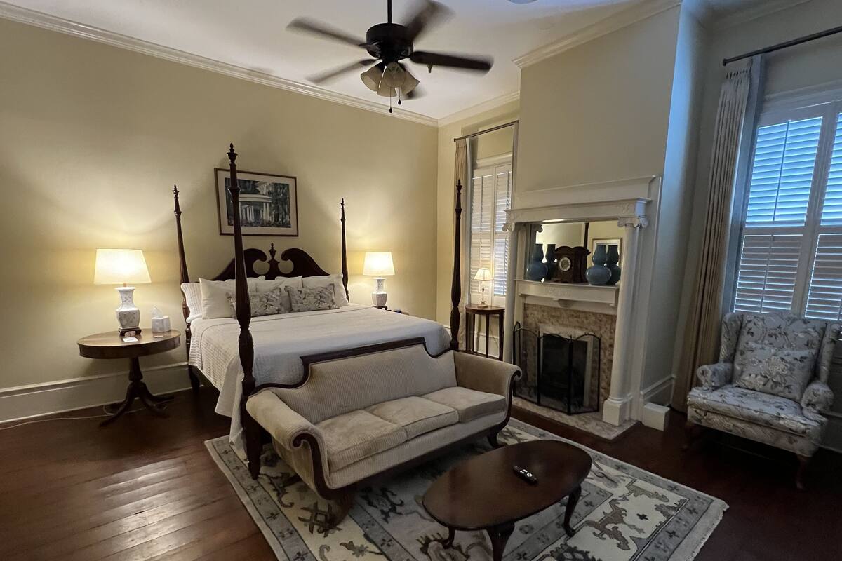 A bedroom with a four-poster bed, two bedside lamps, a fireplace, a sofa, and a chair.