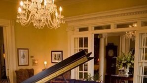 Un piano de cola en una habitación luminosa con una araña, un sofá azul y una alfombra estampada.