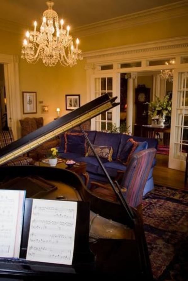A grand piano in a well-lit room with a chandelier, a blue sofa, and a patterned rug.