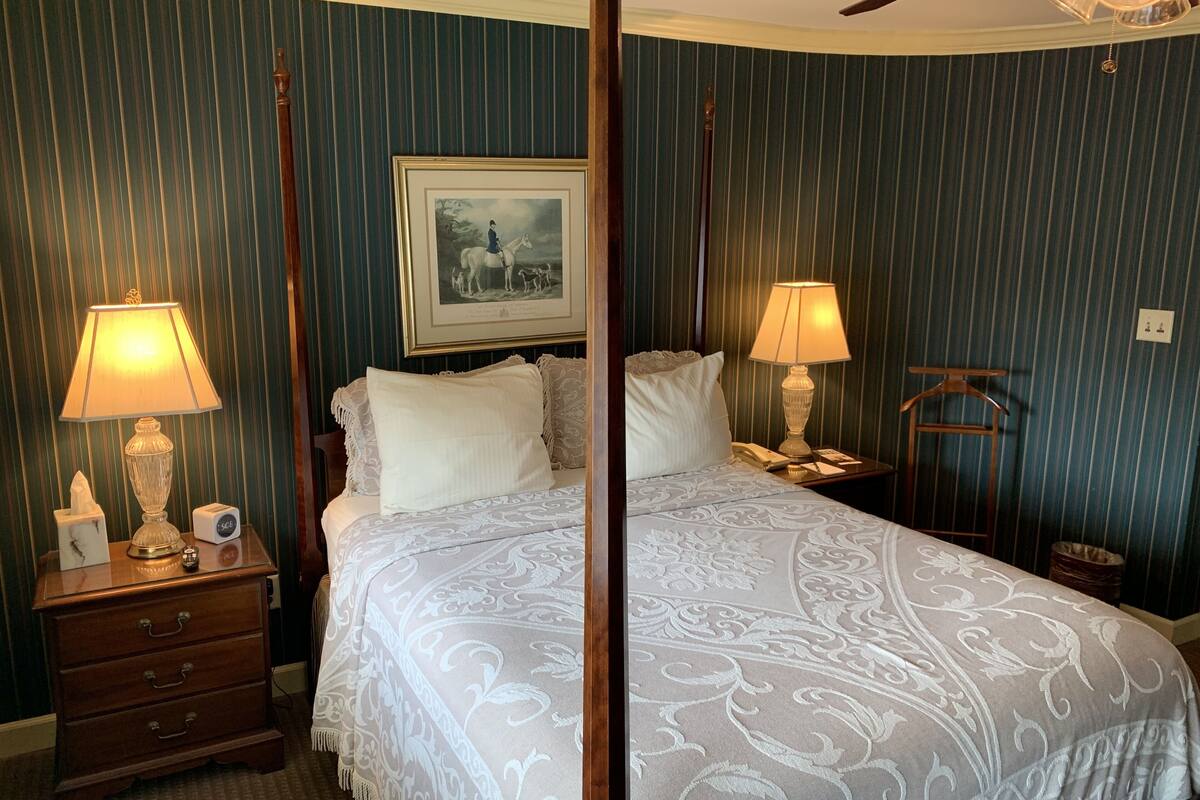 A four-poster bed with a white bedspread, two bedside lamps, a framed picture on the wall, and a wooden nightstand with a clock.
