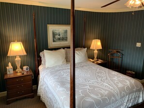 A four-poster bed with a white bedspread, two bedside lamps, a framed picture on the wall, and a wooden nightstand with a clock.