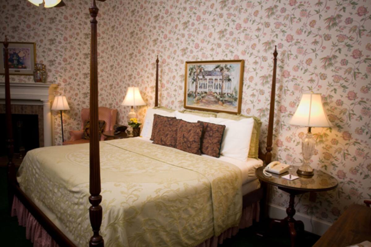A four-poster bed with a floral wallpaper background, a painting above the bed, and two bedside lamps.
