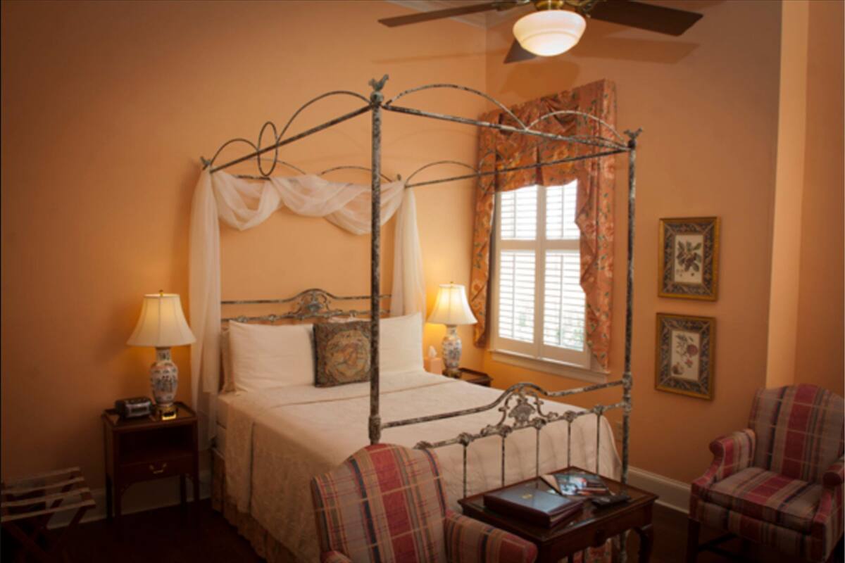 A four-poster bed with a canopy, two bedside lamps, a nightstand, a chair, and a small table with a book.