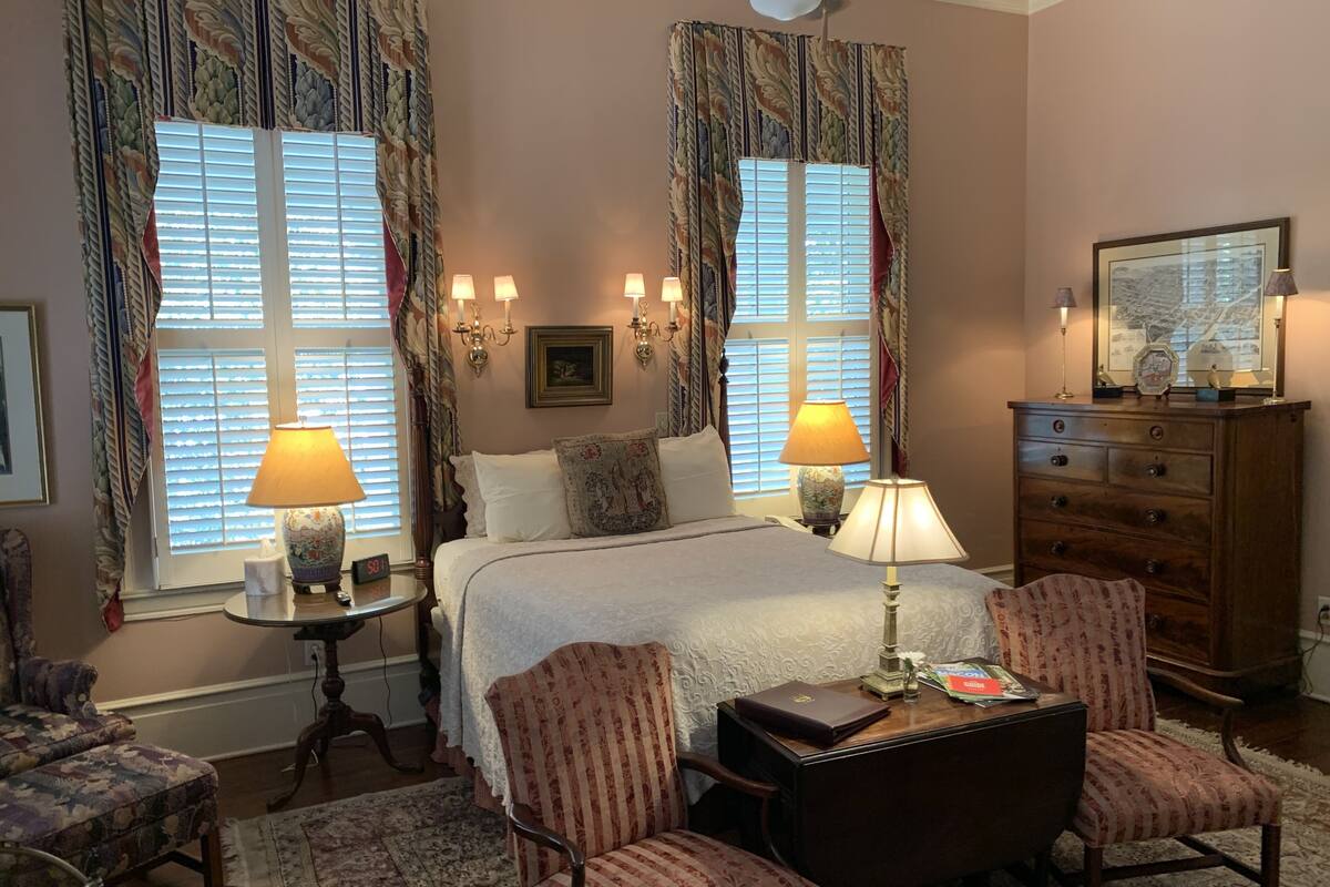 A hotel room with a large bed, two bedside lamps, a wooden dresser, a desk with a chair, and patterned curtains.