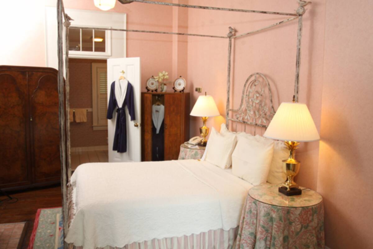 A four-poster bed with white linens, two bedside tables with lamps, a wooden wardrobe, and a mirror on the wall.