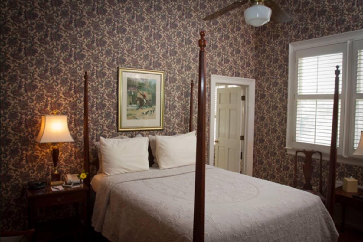 A four-poster bed with a canopy, a nightstand with a lamp, a framed picture on the wall, and a window with blinds.