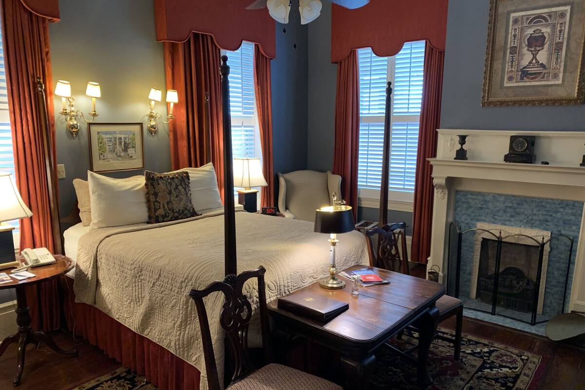 A hotel room with a large bed, a bedside table with a lamp, a chair, a fireplace, and a clock on the mantel.