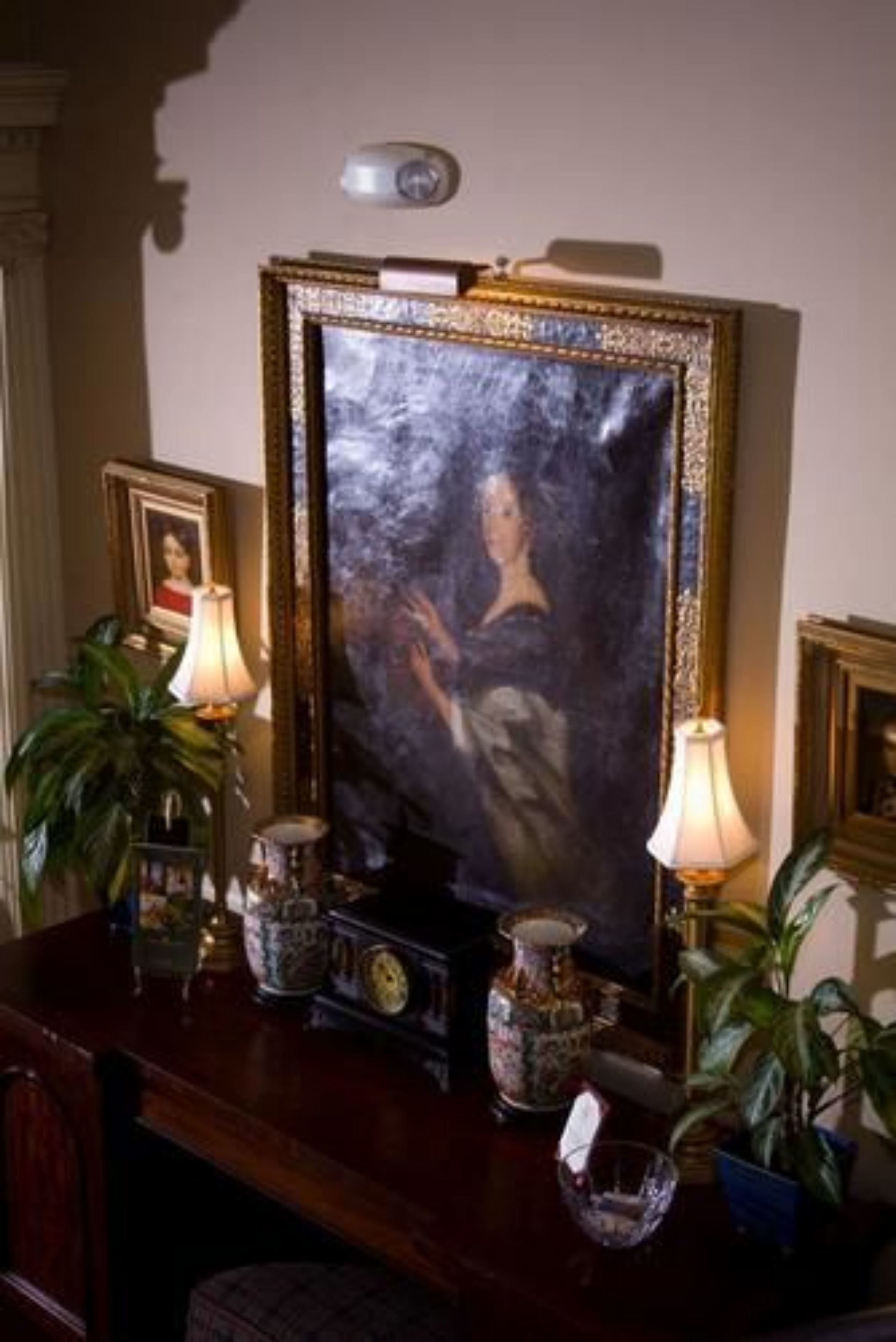 A framed portrait on a wall, a clock, and two lamps on a wooden table.