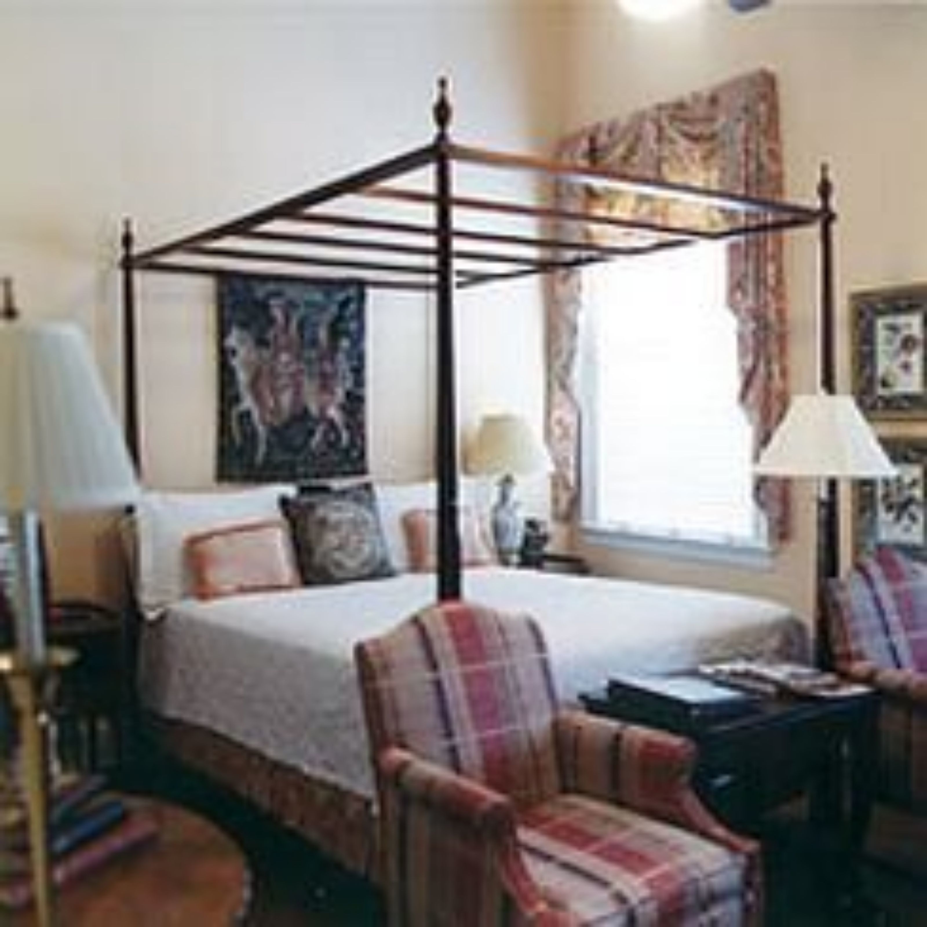 A four-poster bed with a wooden frame, a bedside table with a lamp, a plaid armchair, and a painting on the wall.