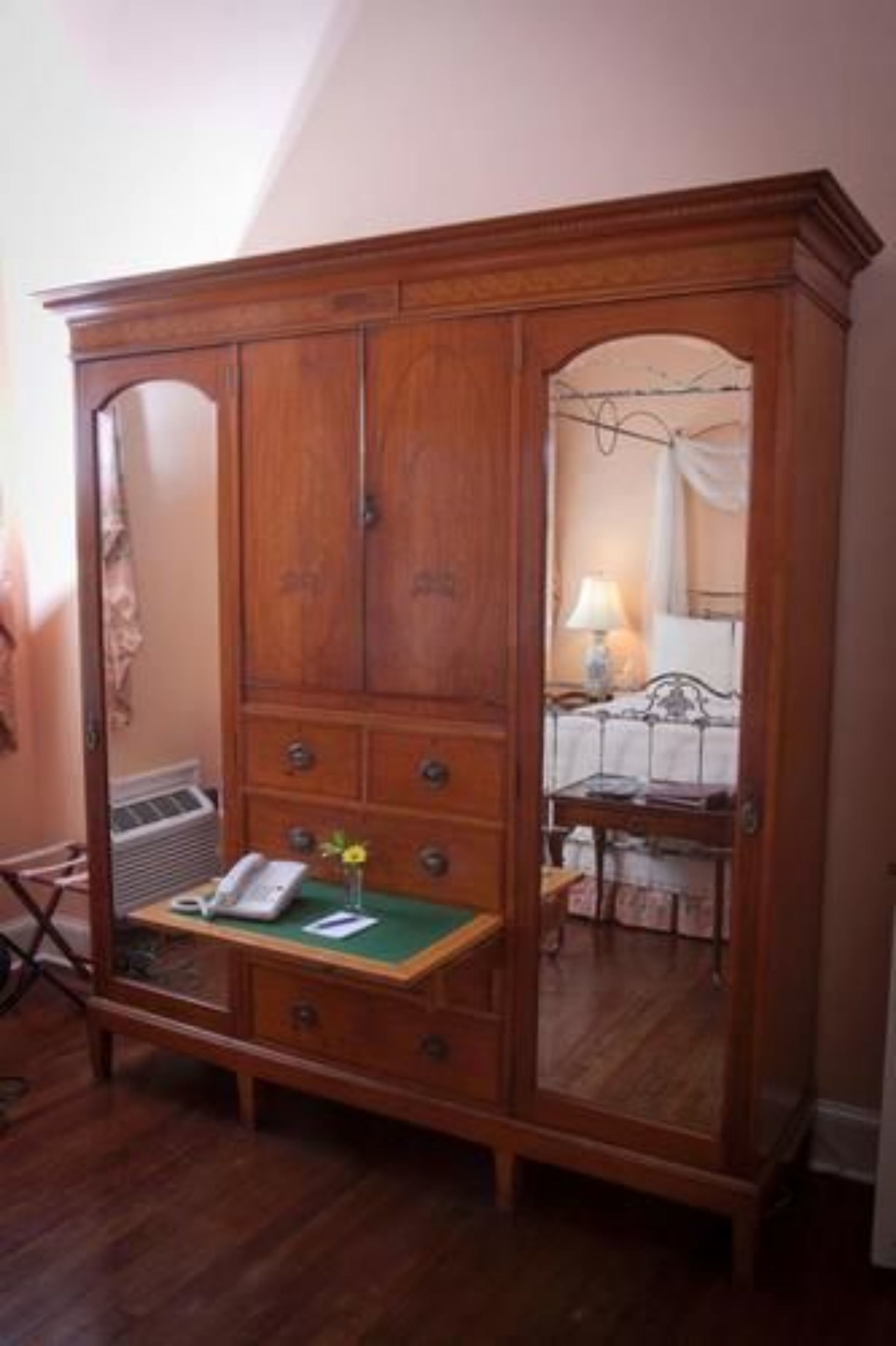 Une armoire en bois avec un miroir, une petite table sur laquelle est posé un vase, et un lit avec une table de chevet en arrière-plan.