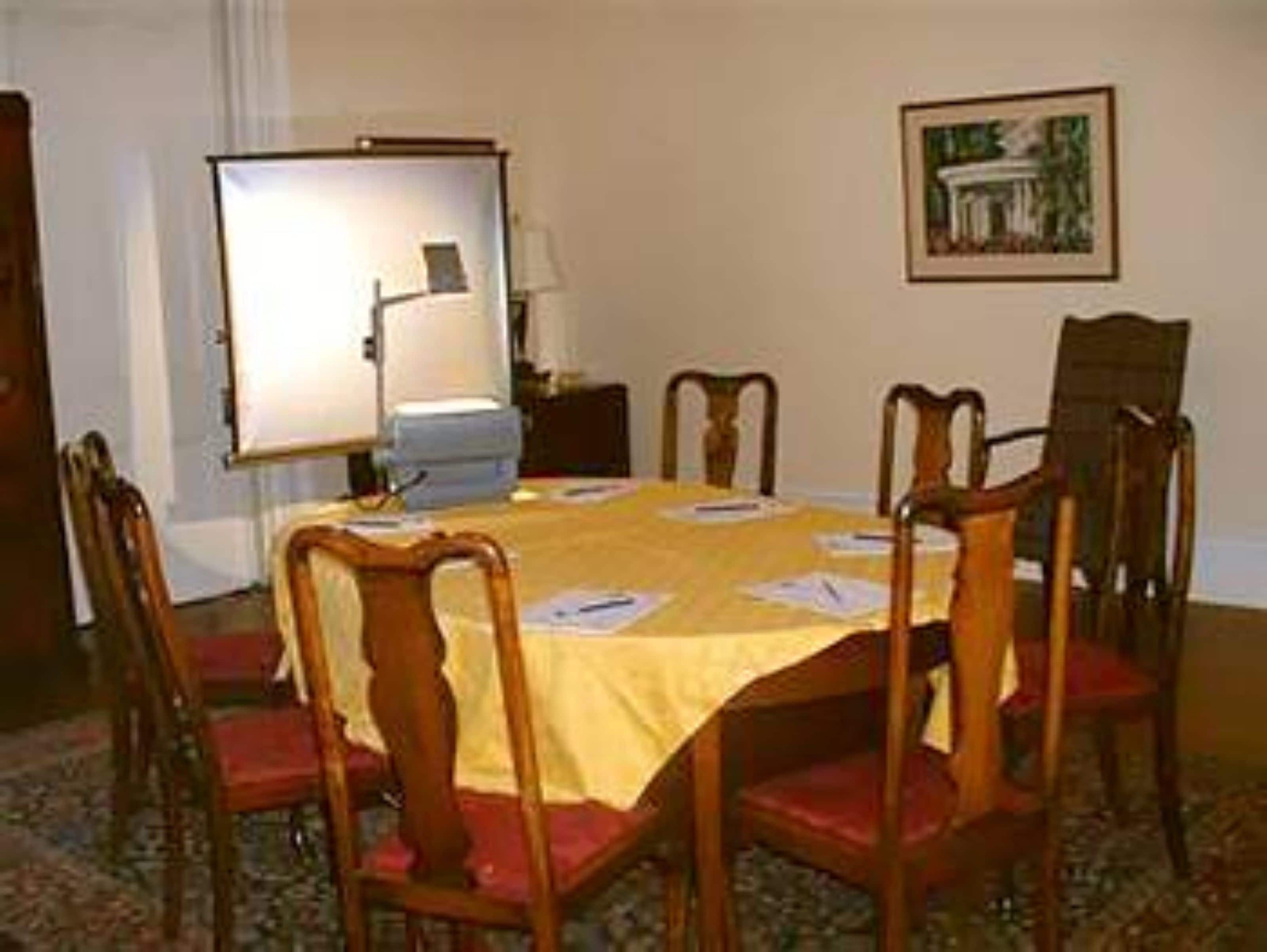 A conference room with a round table, six wooden chairs, a projector screen, and a framed picture on the wall.