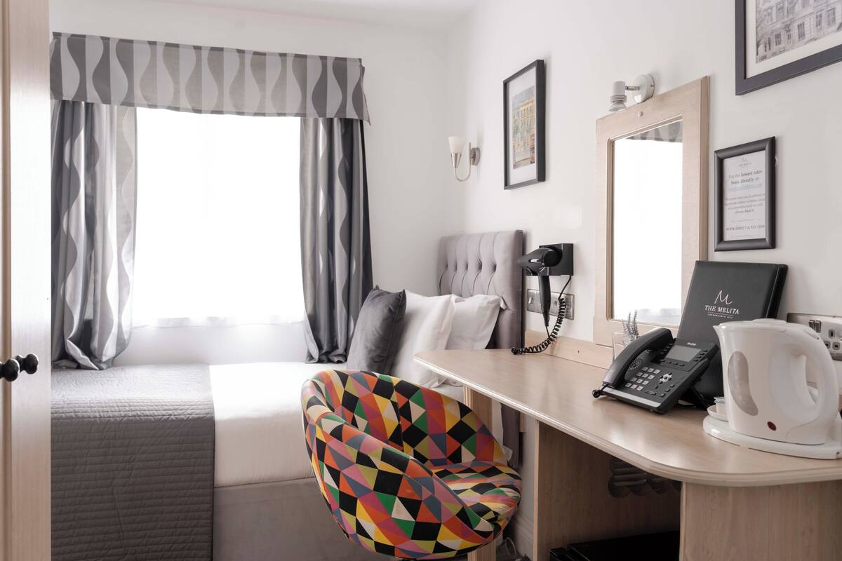 A hotel room with a bed, a desk with a telephone and a coffee maker, a colorful chair, and a window with curtains.